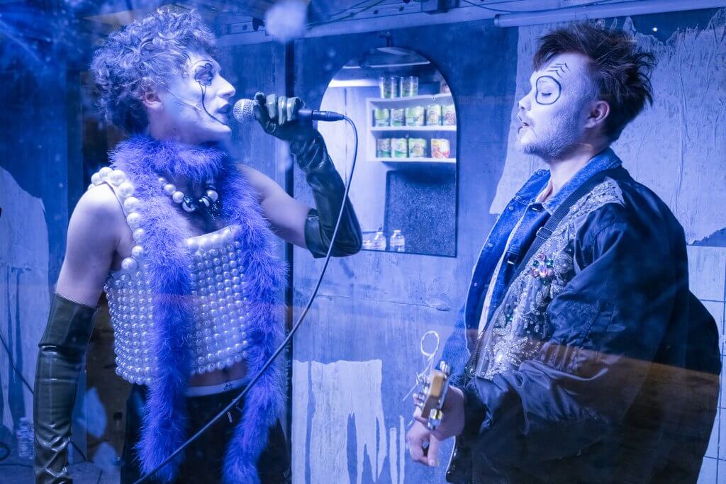 Zwei Personen mit clownesk geschminkten Gesichter stehen sich auf der blau erleuchteten Bühen gegenüber, der eine singt in ein Mikro, der andere spielt Gitarre.