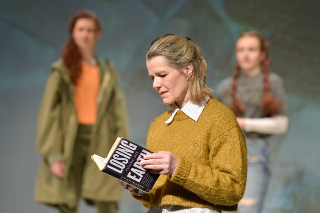 Eine Frau in senfgelben Pullover blättert in einem Buch, unscharf dahinter sind zwei Frauen zu sehen.