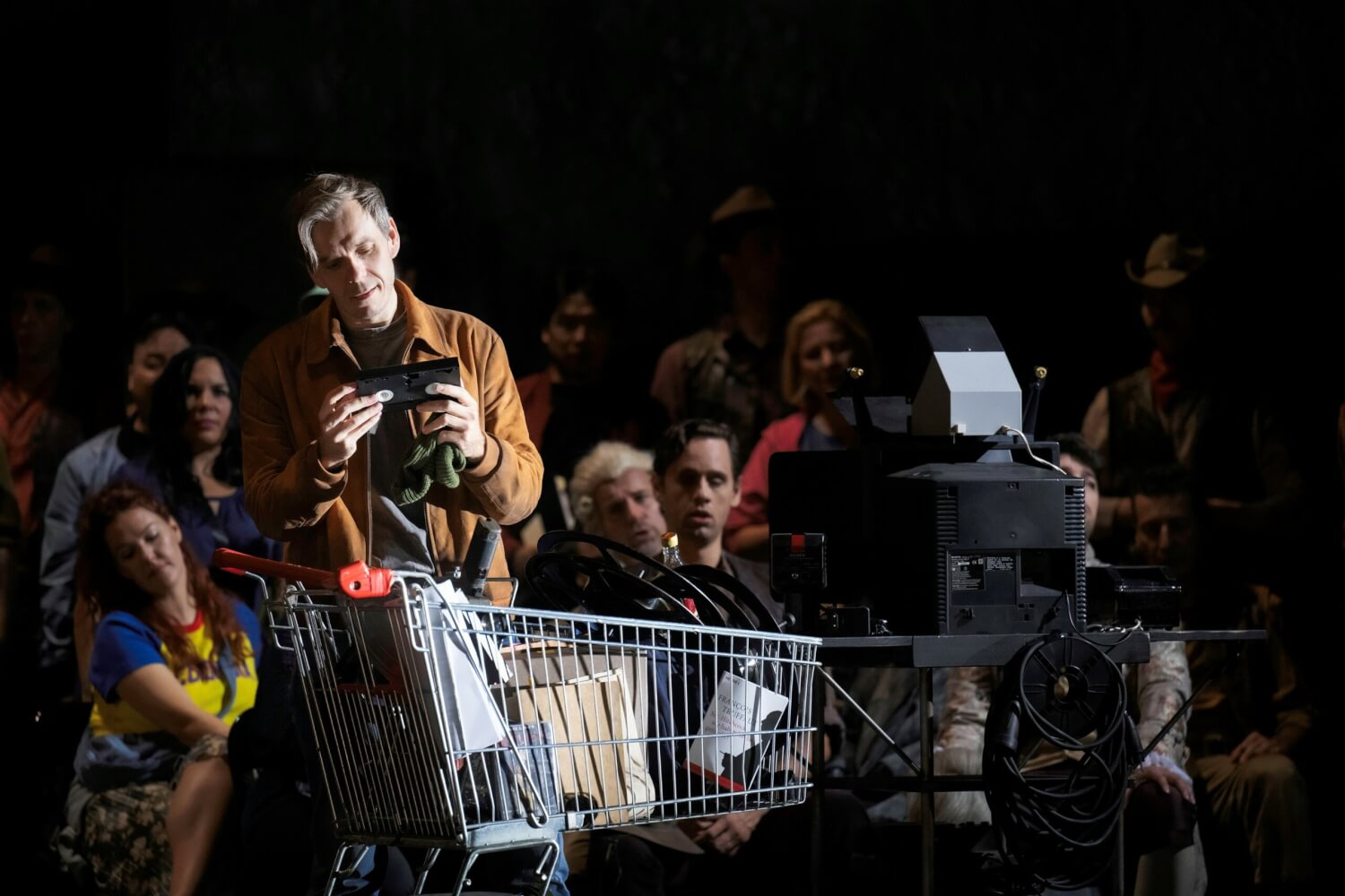 Benjamin Bernheim (Hoffmann) steht vor einer Menschenmenge (Chor) mit einem Einkaufswagen. Er hält eine Videokassette in den Händen.