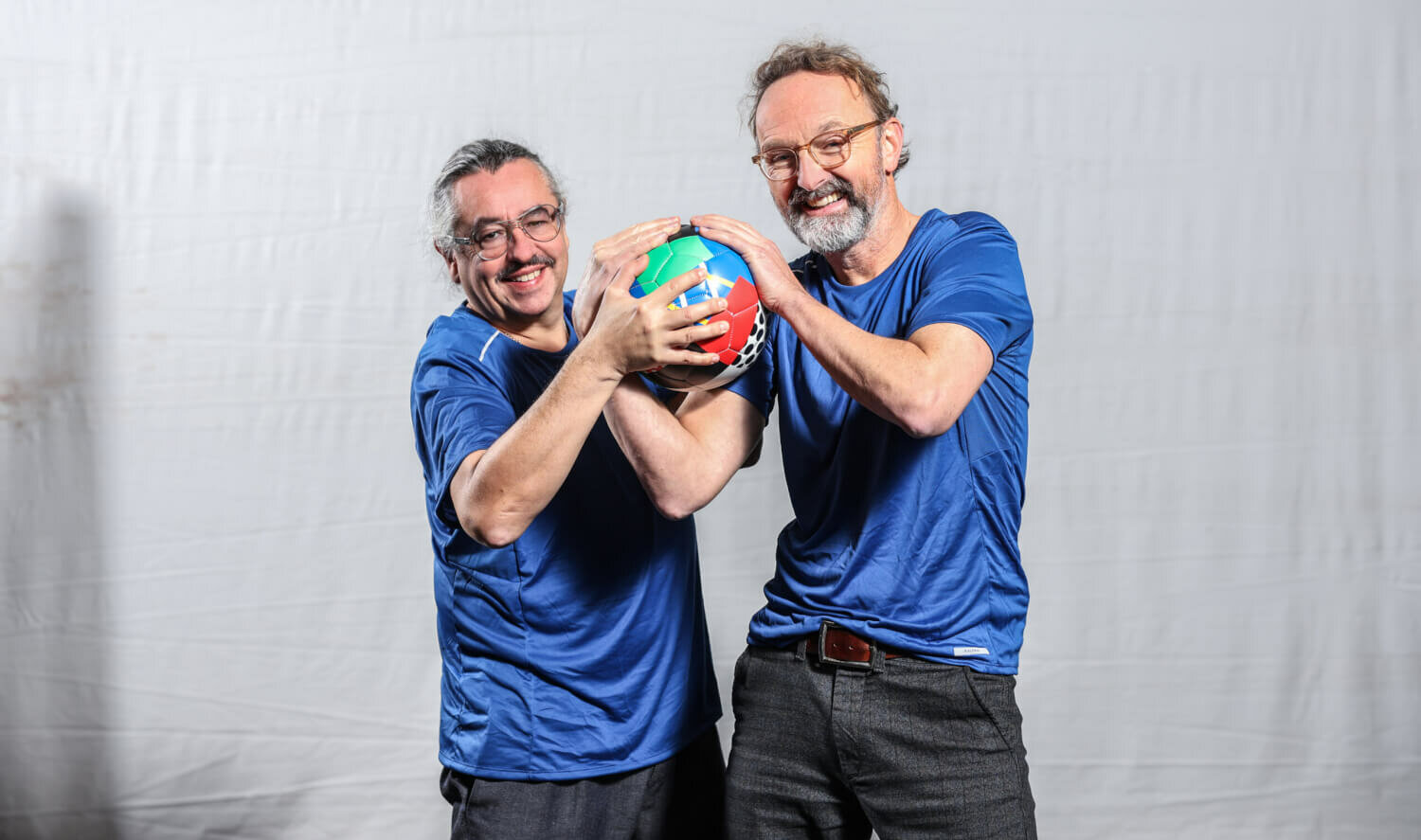 Zu sehen sind Leonhard Koppelmann und Peter Jordan, beide haben blaue T-Shirts an und umklammen gemeinsam einen bunten Handball